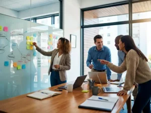 Diverse professionals collaboreren rond whiteboard met kleurrijke sticky notes, persoon plaatst gele notitie terwijl anderen betrokken toekijken in modern kantoor